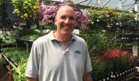 Smiling man in a gray polo shirt standing among vibrant flowers and plants in a greenhouse, representing landscape care expertise for Scape-abilities.