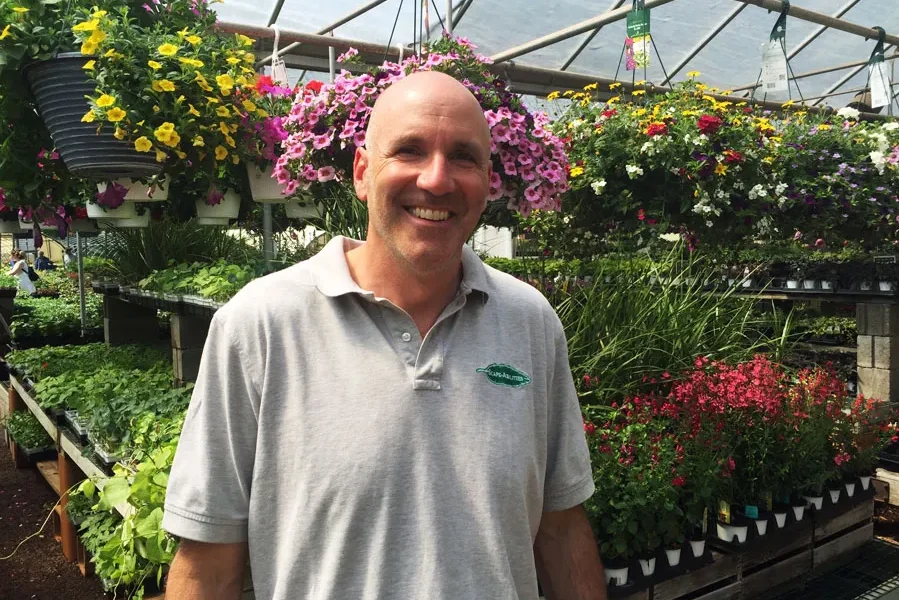 Stephen Costalos smiling in a greenhouse surrounded by vibrant flowers and plants, representing expertise in landscape design and lawn care services offered by Scape-abilities.